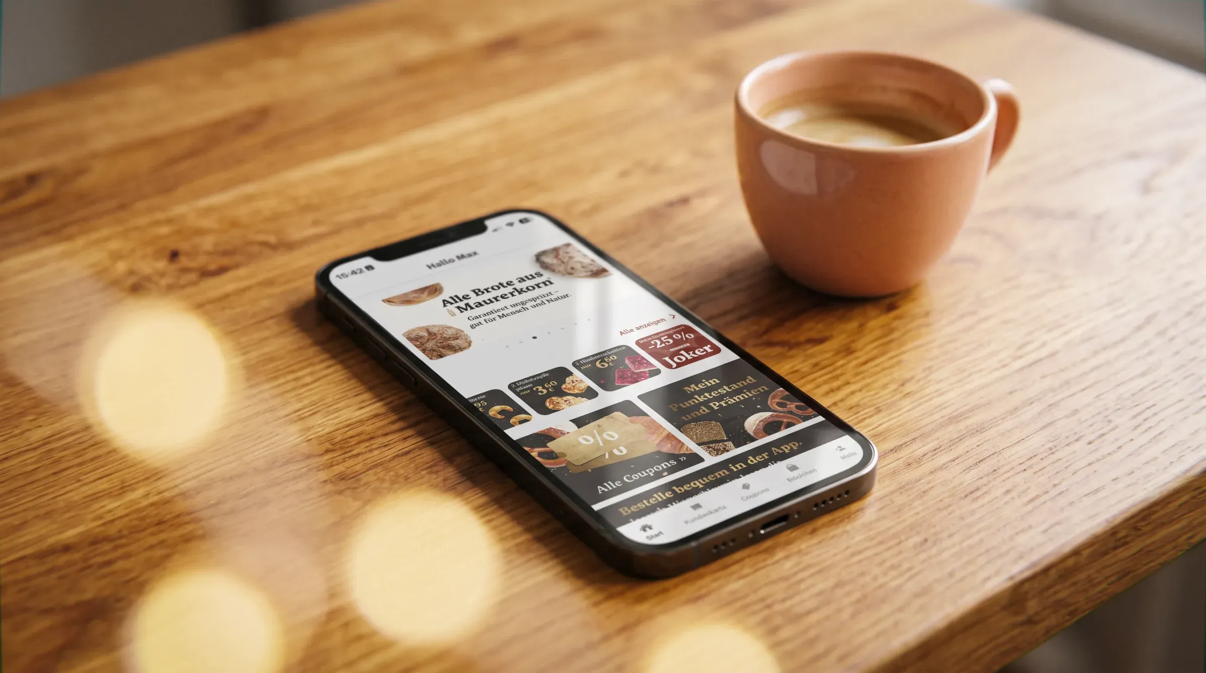 Ein Telefon liegt auf einem Holztisch neben einer Tasse, auf dem Bildschirm die Startseite der Maurer-App mit den Broten aus Maurerkorn
