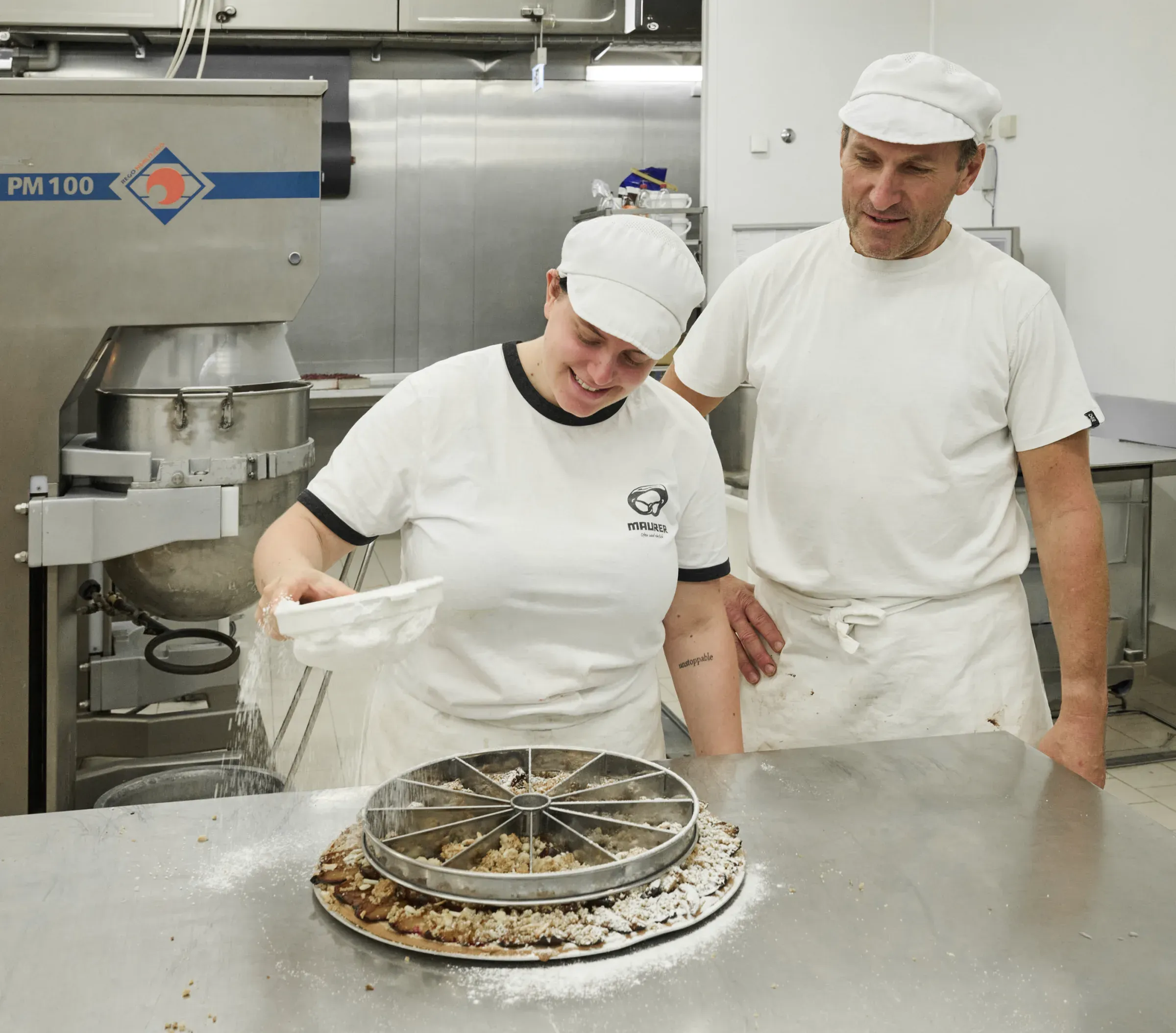 Mitarbeiterin bestäubt einen Streuselkuchen in der Form mit Puderzucker, ein Kollege sieht zu