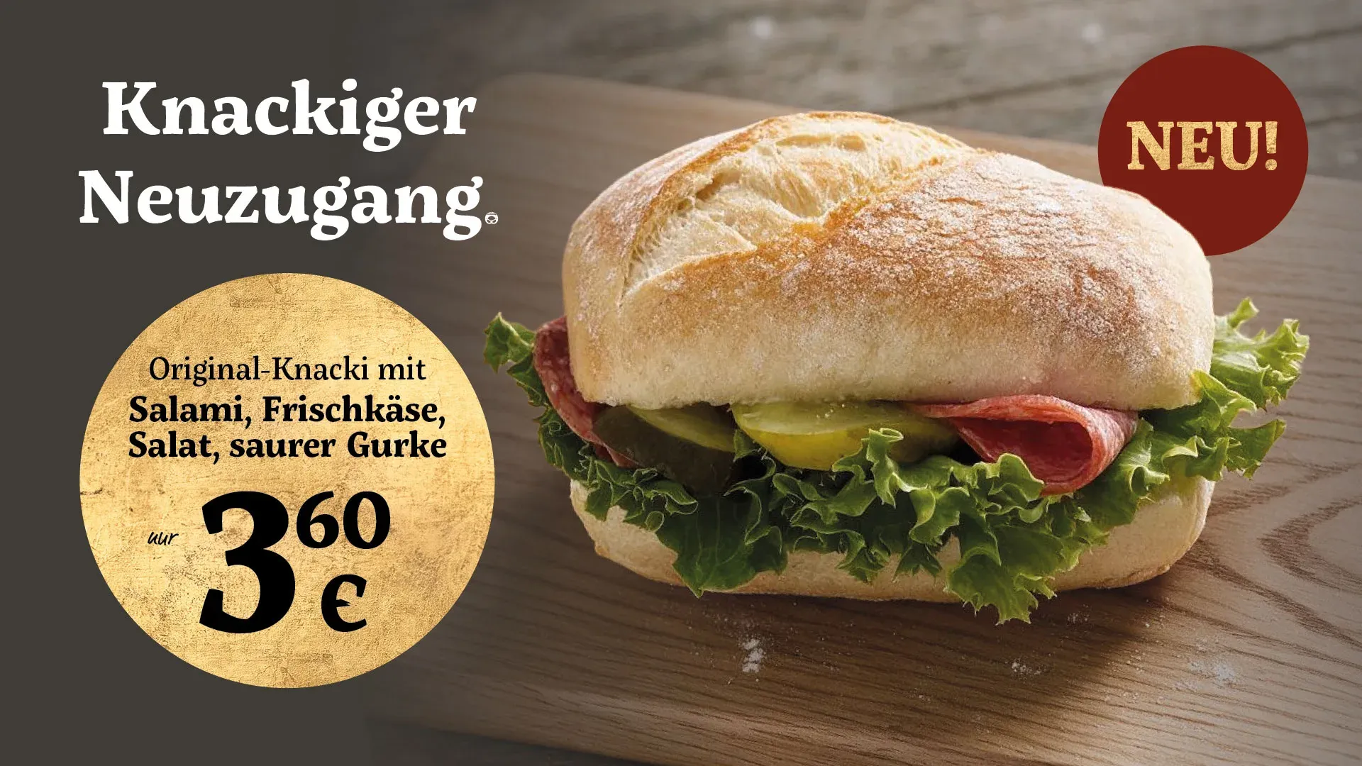 Aushang: ein belegter Knacki auf einem Holzbrett. Darauf steht „Knackiger Neuzugang“, „Neu!“ und „Original-Knacki mit Salami, Frischkäse, Salat, saurer Gurke, nur 3,60 €“