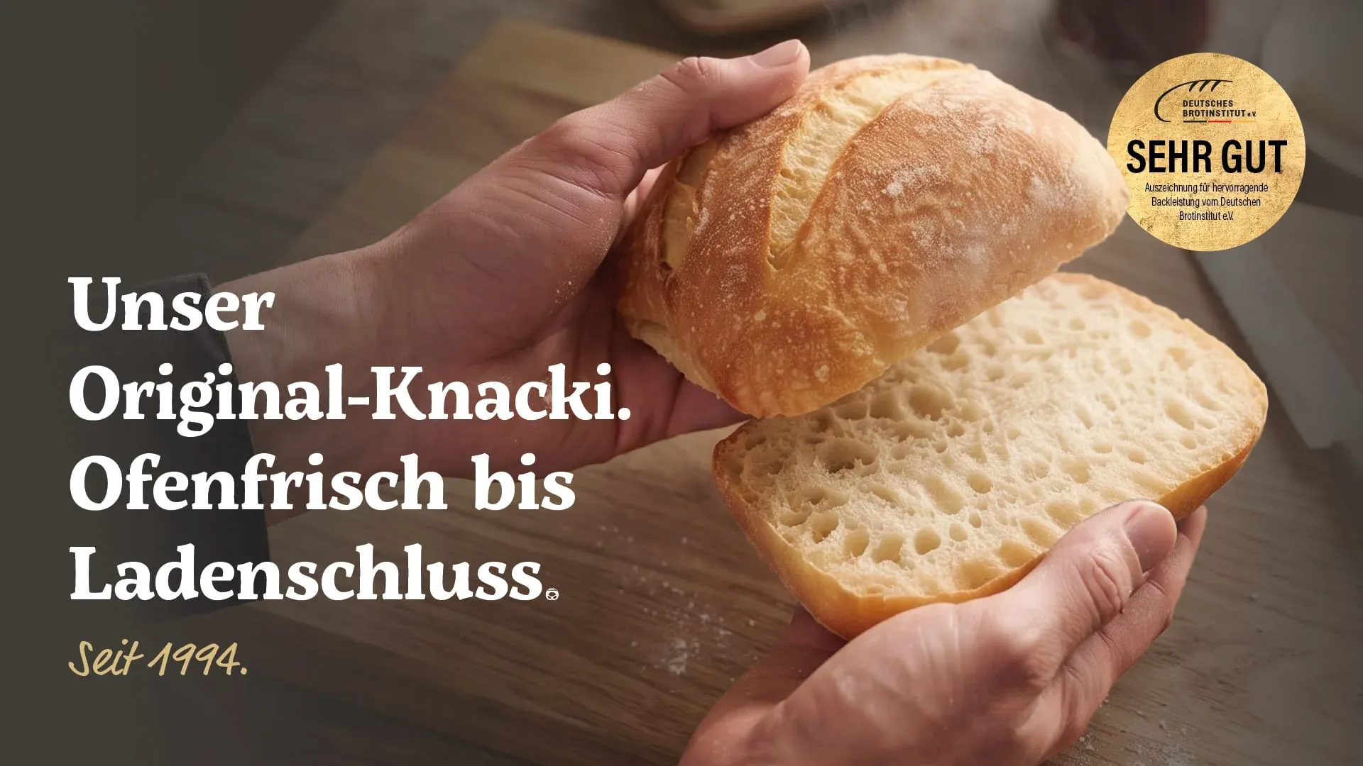 Aushang: zwei Hände halten einen aufgeschnittenen Knacki. Darauf steht „Unser Original-Knacki. Ofenfrisch bis Ladenschluss. Seit 1994“ und das Siegel „sehr gut“ des Deutschen Brotinstituts