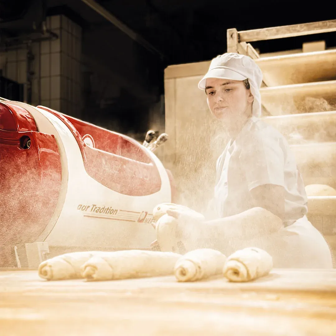 Bäckerin arbeitet im Mehlstaub an der Teigmaschine, vor ihr liegen aufgerollte Teiglinge