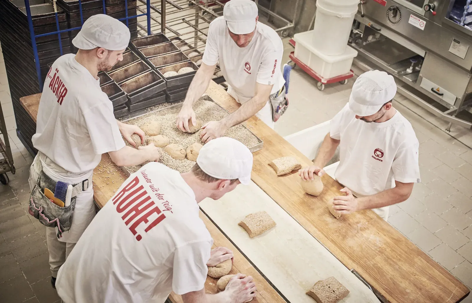 Vier Bäcker formen gemeinsam Brote an einem langen Arbeitstisch in der Backstube