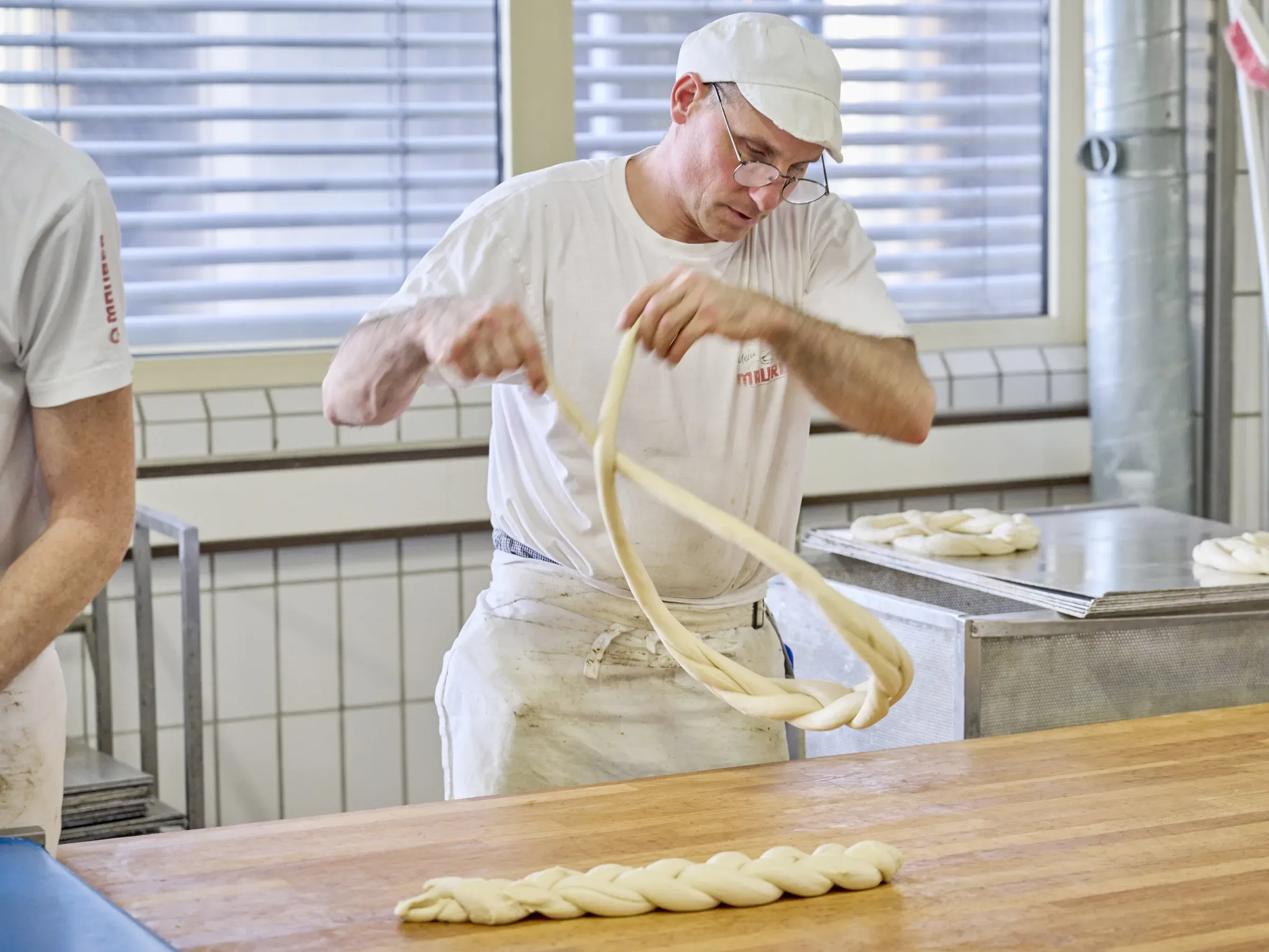 Bernd Simon schlingt einen langen Hefeteigstrang zu einer Neujahrsbrezel, vor ihm liegt eine fertig geschlungene auf dem Holztisch