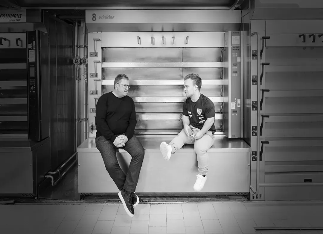 Tobias Maurer und Niko Kappel im Gespräch auf der Ofenbank zwischen den Öfen der Backstube, schwarzweiß