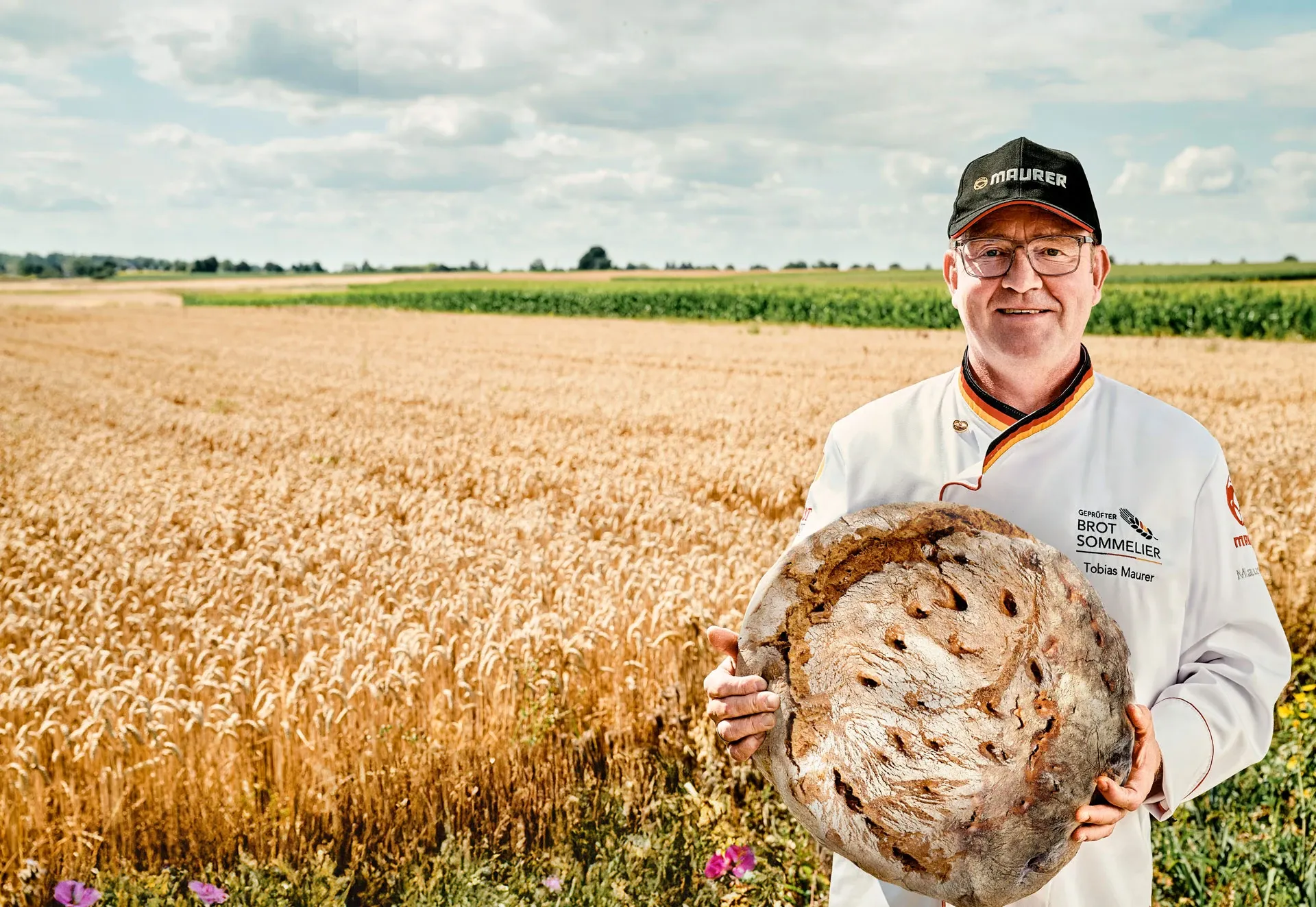 Tobias Maurer in der Sommelier-Jacke vor einem reifen Kornfeld, in den Händen ein großer Laib