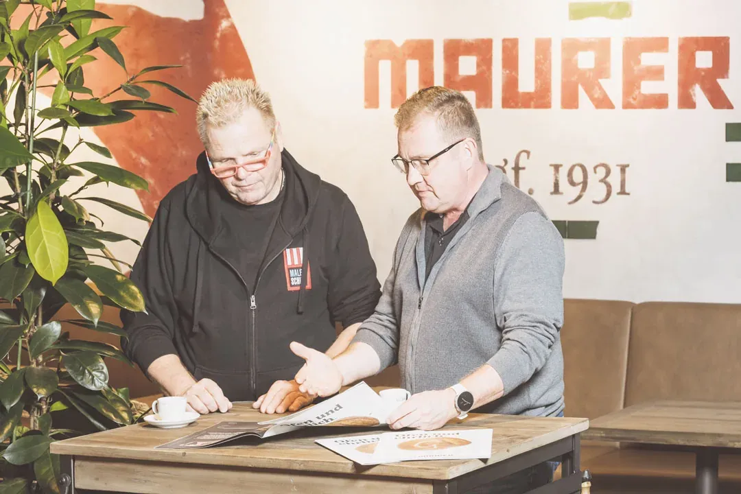 Kurt Schief und Tobias Maurer stehen an einem Tisch über einem aufgeschlagenen Magazin, dahinter die Wand mit dem Schriftzug MAURER ågf. 1931