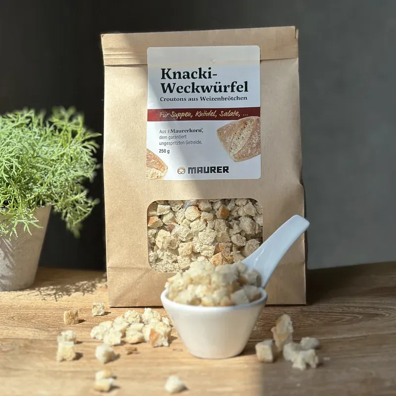 Papiertüte Knacki-Weckwürfel neben einer Schale Croutons mit Porzellanlöffel auf einem Holztisch